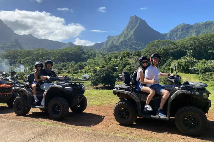 People on ATVs posing with a mountain and forested landscape in the background, under a clear blue sky.