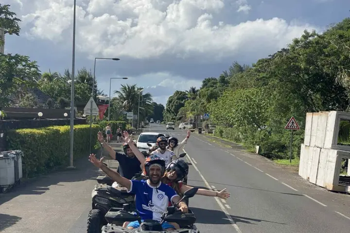 Group of people happily riding quad bikes on a sunny road with lush greenery and cloudy sky.