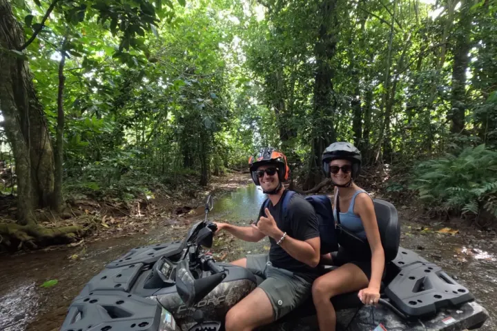 Two people on an ATV in a forested area with helmets and sunglasses.