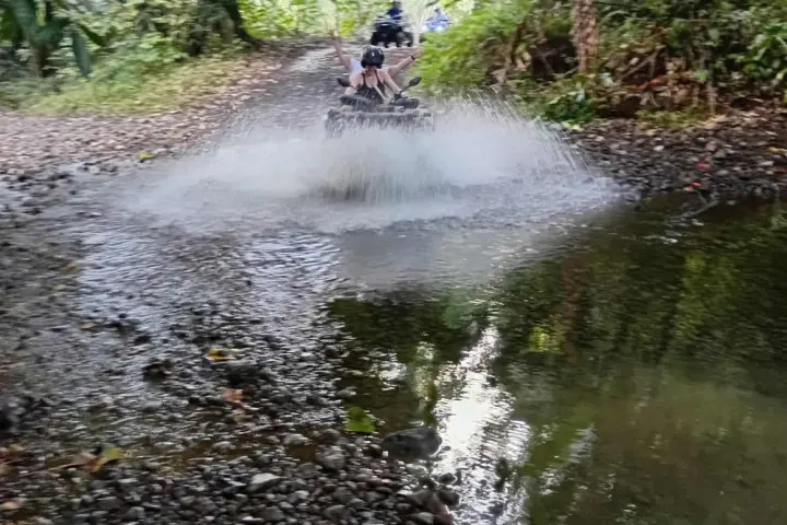 Person riding an ATV through a shallow stream on a forest trail, splashing water.