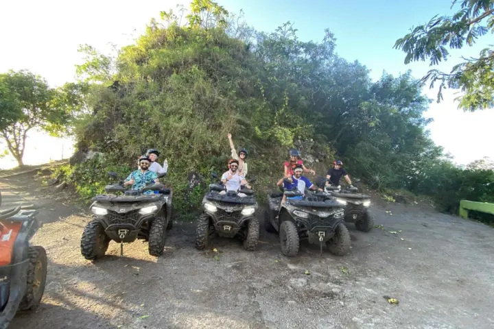Group of six people on ATVs near a verdant hill, smiling and posing for the camera.