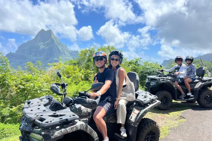 Two couples on ATVs on a scenic road with mountains and blue sky in the background.