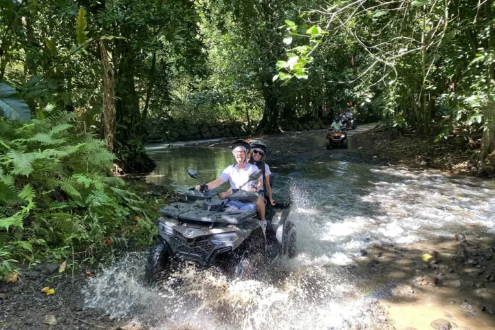 Two people riding an ATV through a shallow river in a forested area.