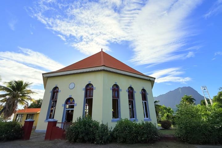 Octagonal building with red roof, arched windows, and lush plants against a blue sky.