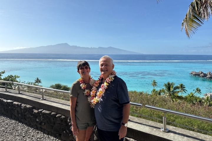 Two people wearing leis stand by a tropical ocean with mountains and palm trees in the background.