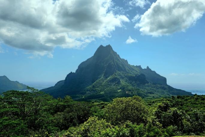Mountain with lush greenery and ocean in the background under a blue sky with clouds.