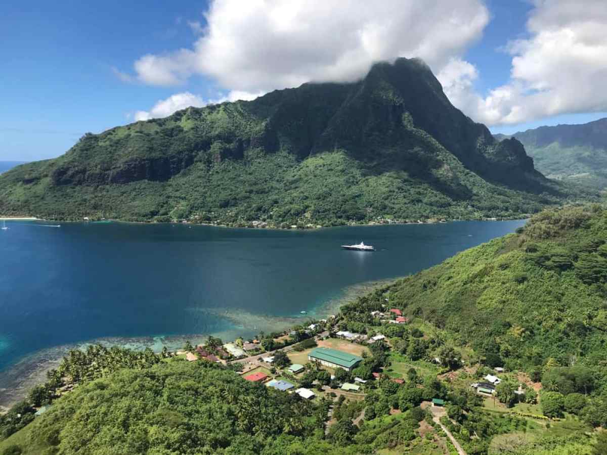 Lush green mountains surround a blue bay with a yacht and scattered buildings.
