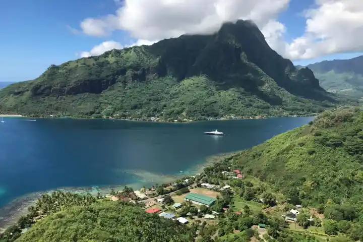 Lush green mountains surround a blue bay with a yacht and scattered buildings.